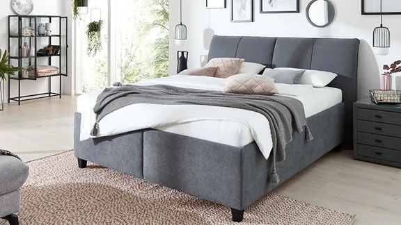 Ein dunkelgraues Polsterbett mit weißer Bettwäsche und grauer Tagesdecke in einem modernen, hellen Schlafzimmer.