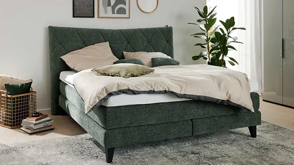 Ein dunkelgrünes Boxspringbett mit Rautenheftung und beiger Bettwäsche, arrangiert in einem hellen Schlafzimmer.